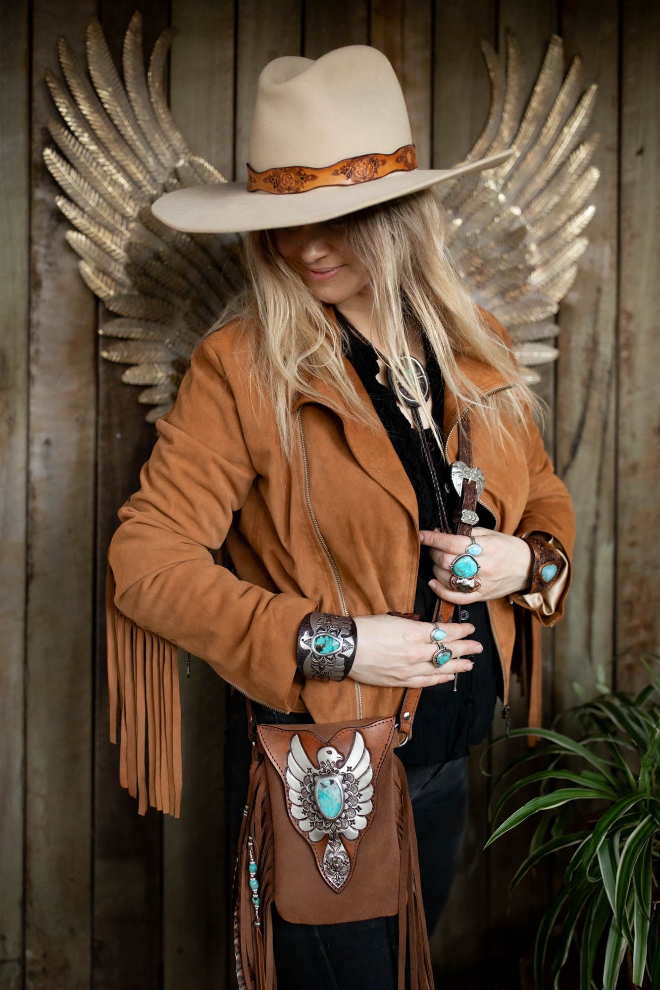 Person wearing a tan suede jacket with fringe, white cowboy hat, and turquoise jewelry against a wooden wall.
